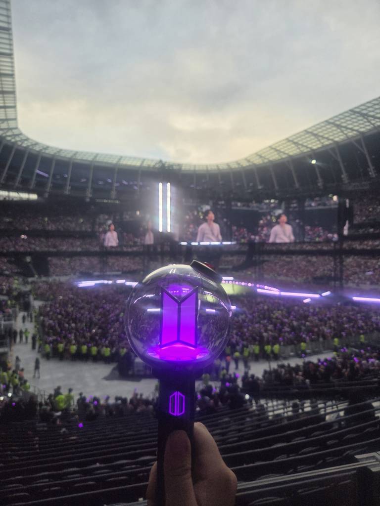 An ARMY BOMB / BTS Lightstick glowing purple, held up in the foreground. In the background is a crowd of BTS fans, and Min Yoongi projected on a large screen as he performs on stage.